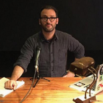 Middle aged white man with dark hair, beard and dark rimmed glasses with black shirt sitting at wooden table with a tabletop microphone in front of him and a desk lamp and a computer and papers