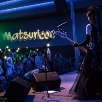 White woman in a Renaissance looking gown playing guitar on stage to a crowd standing under the sign Matsuricon