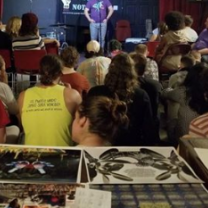 Lots of people with their backs to the camera sitting and watching someone standing at the mic, with art on a table in the foreground