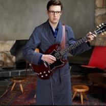 Young geeky guy with big black rimmed glasses wearing a bathrobe holding an electric guitar in a room with two wooden stools