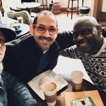 A balding white guy with red framed glasses and facial hair smiling sitting at a table with his arm around a bald black man in a white and black designed sweater also smiling