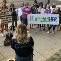 A back of a woman in the foreground speaking outside to a group of women facing her holding signs saying #Fight4Her