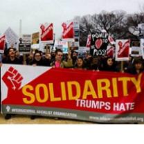 Lots of people at a march holding picket signs and a very large long banner with red fist and words Solidarity trumps hate