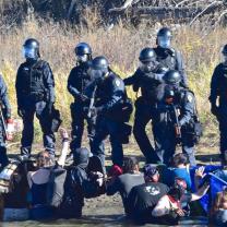 Photo of heavily armed soldier-looking men pointing guns at Native American protestors sitting on the ground
