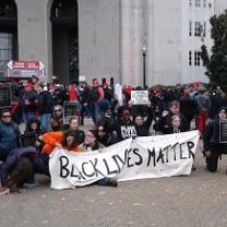 People wearing winter clothes bending down on one knee outside holding a long white banner with black letters reading Black Lives Matter in front of a large white building