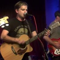 White man with brown hair playing a guitar and singing at a mic with other musicians