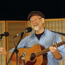 Man with gray goatee and black cap holding guitar at microphone smiling 