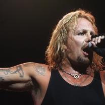 Vice Neil singing into mic with long stringy blonde hair and lots of tattoos