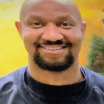 Bald black man with dark mustache and goatee, wearing blue shirt smiling