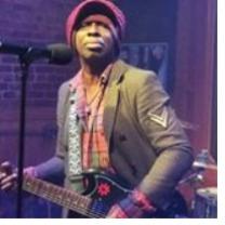 Black man in a hat playing a guitar at a mic