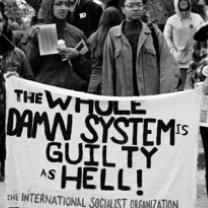 Black and white photo with people in winter coats outside gathered in a group behind a white banner with black letters saying The Whole Damn System is Guilty as Hell